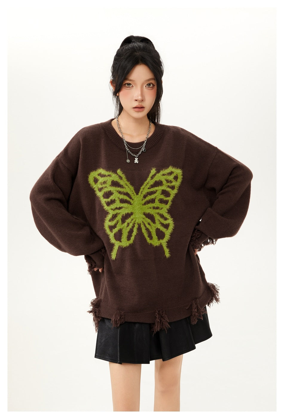 Close three-quarter view of model in brown sweater with green fuzzy butterfly, fringe cuffs and hem, black skirt