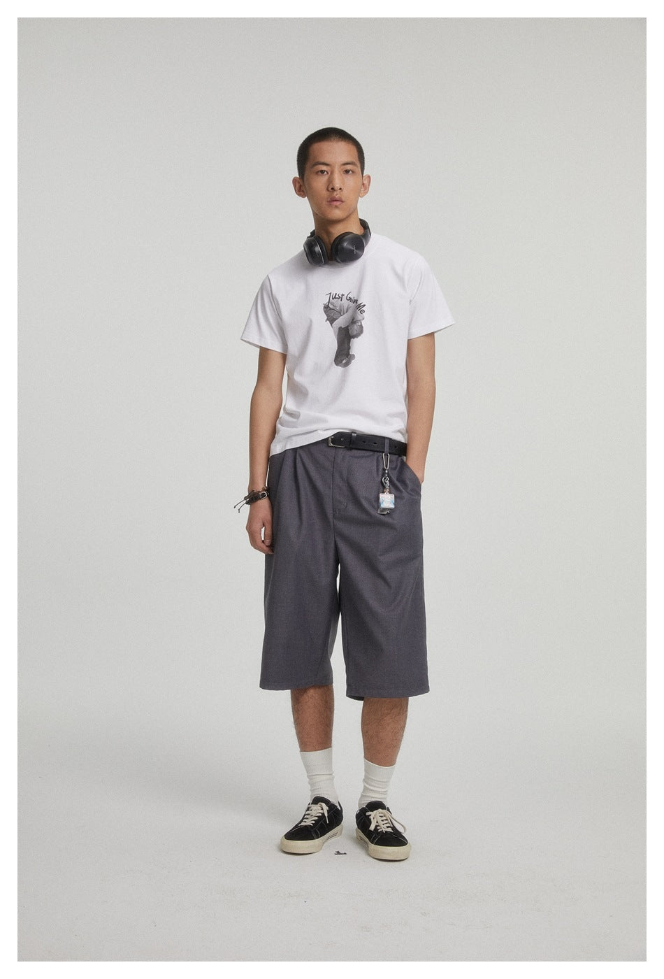 Model wearing a white graphic T-shirt and loose charcoal shorts, shown with headphones around his neck.