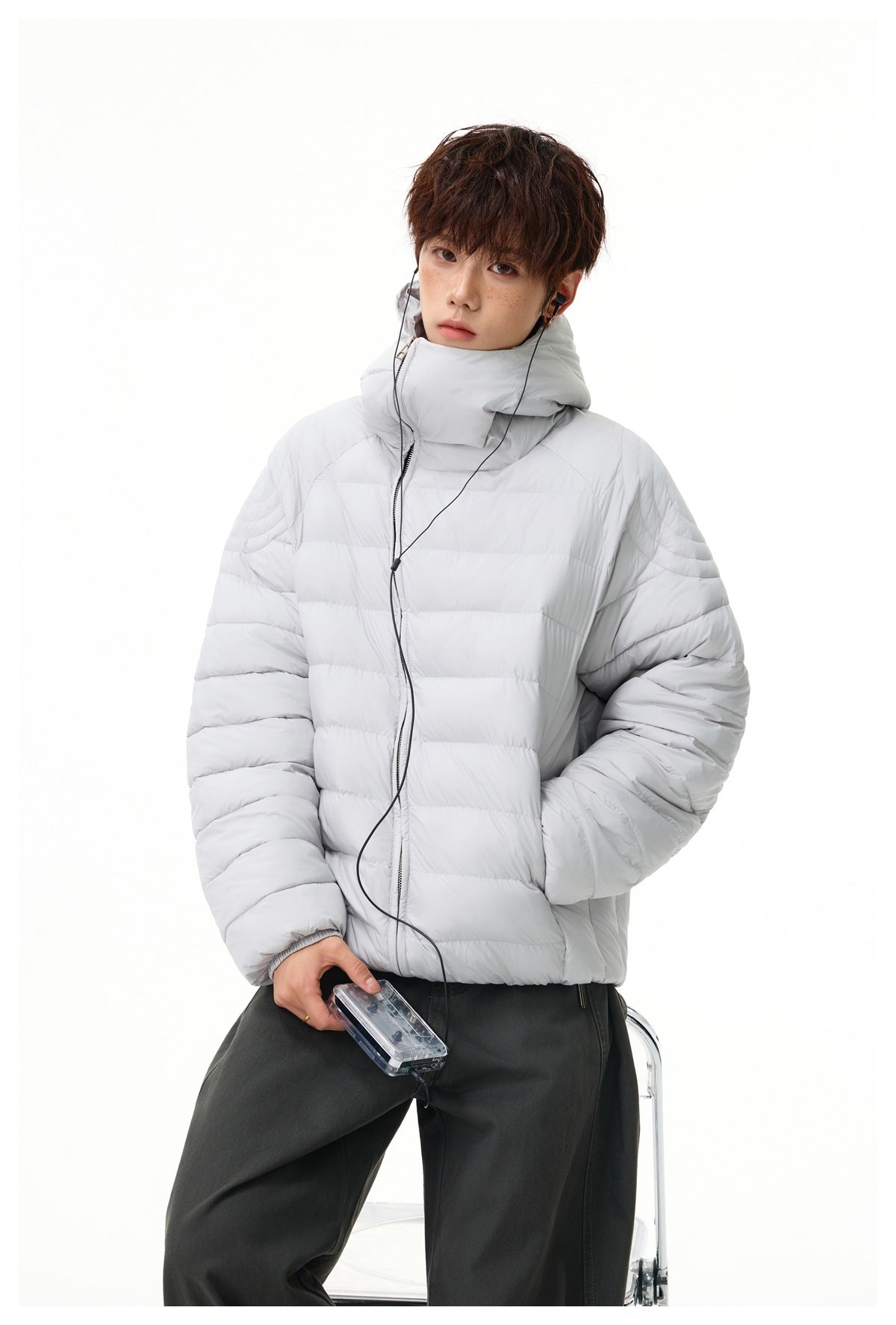 Model wearing light gray hooded quilted jacket with dark gray pants, holding a clear cassette player