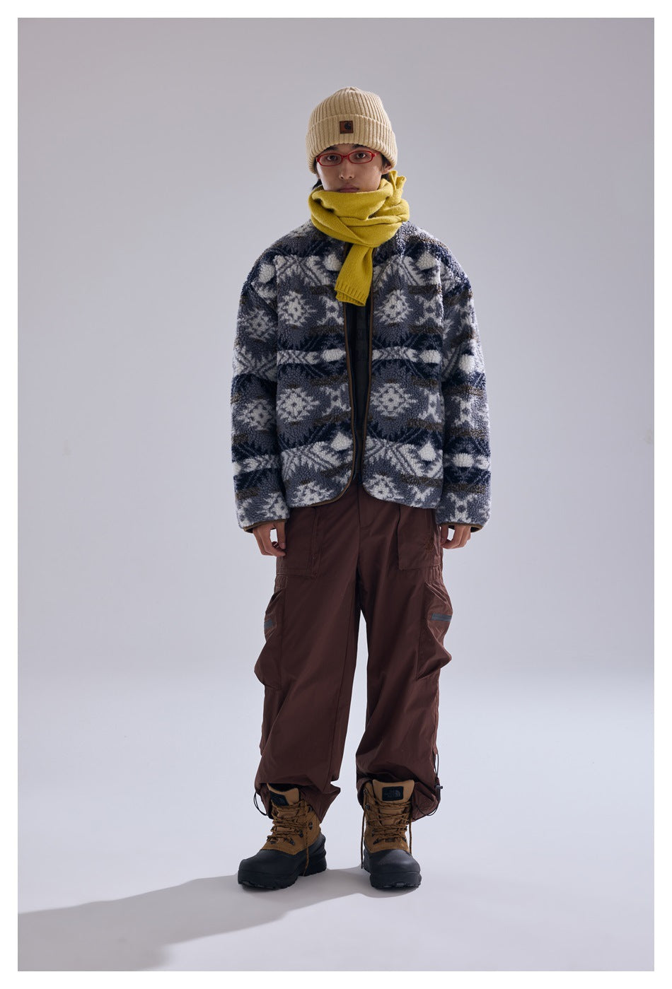 Model wearing gray geometric pattern fleece-like zip jacket with yellow scarf, beanie, brown cargo pants and boots, front