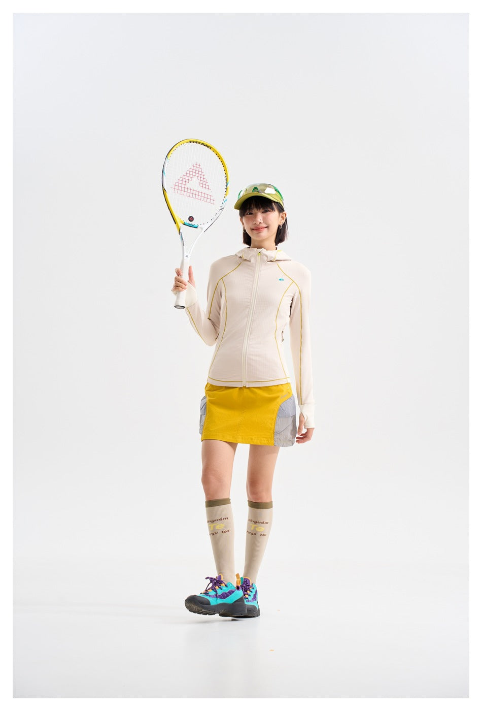 Model holding a tennis racket and wearing a cream hooded jacket with yellow piping and a yellow-and-gray skirt.