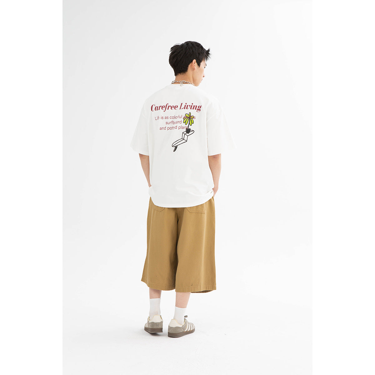 Back view of a model wearing a white T-shirt with colorful flower and surfboard artwork and tan shorts.