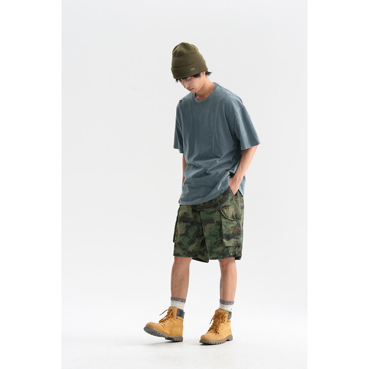 Man wearing an oversized slate blue T-shirt and dark green camouflage cargo shorts against a white studio background.