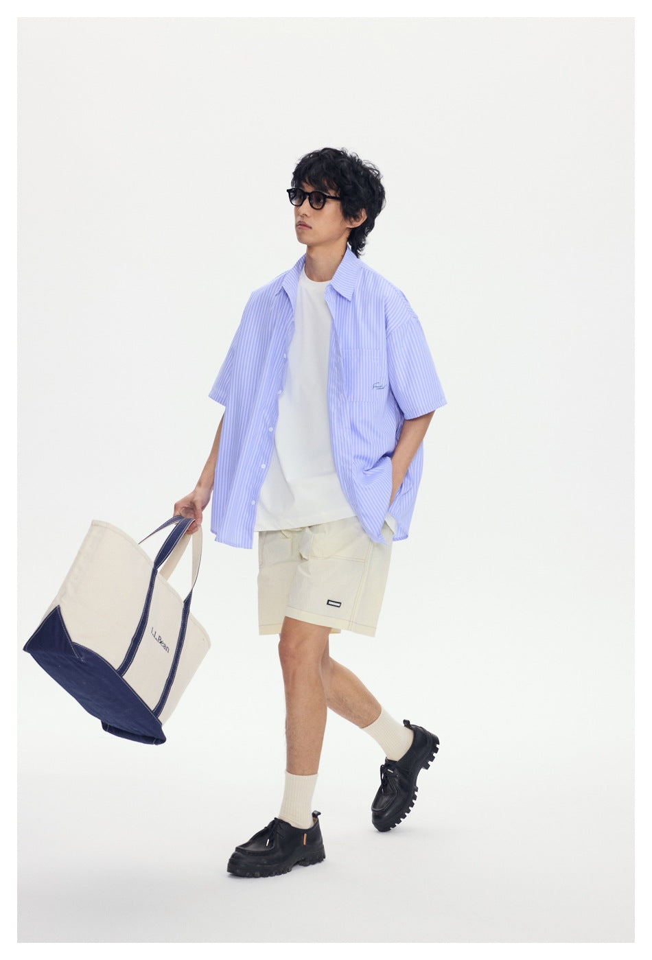 Model walking in an oversized light blue-and-white striped short-sleeve shirt, white tee, and cream shorts.
