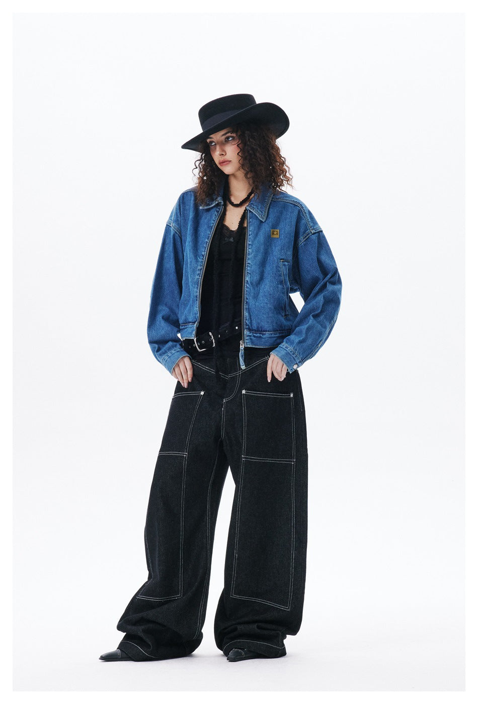 Model wearing cropped blue denim zip jacket open over a black top with black wide-leg jeans and a black hat