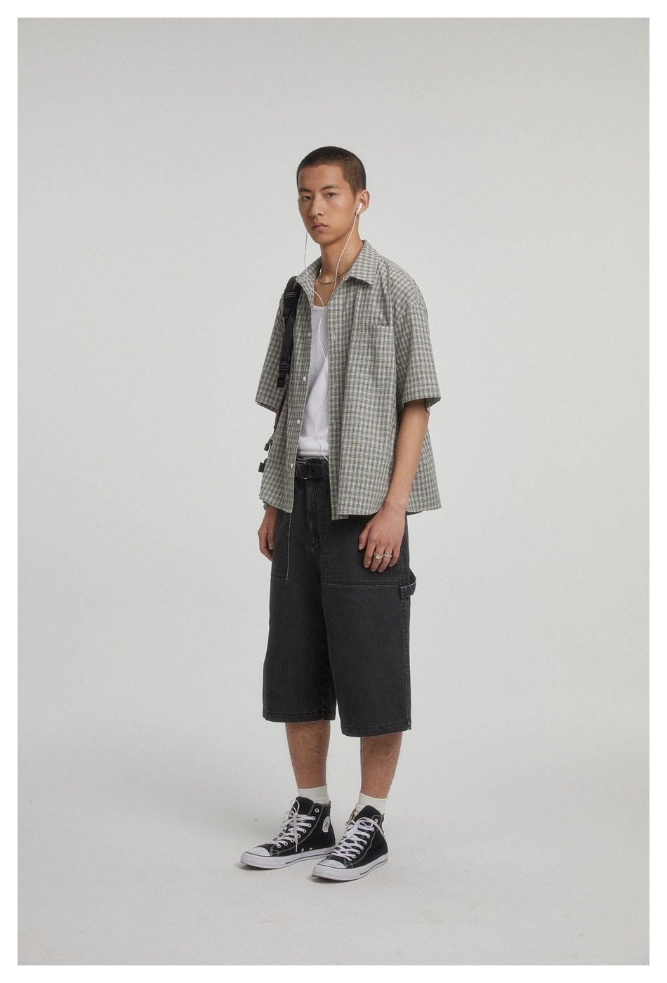Model wearing an open gray checked short-sleeve shirt over a white tank top, with a shoulder bag and dark shorts.