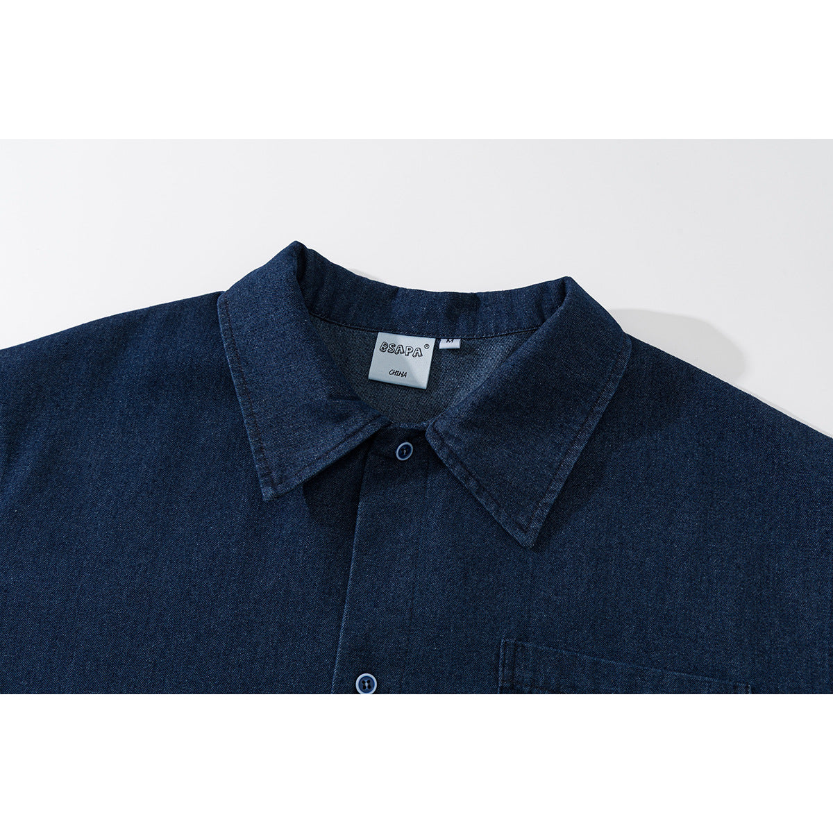 Close-up of a dark indigo button-up shirt collar, buttons and chest pocket; neck label reads BSAPA and CHINA.