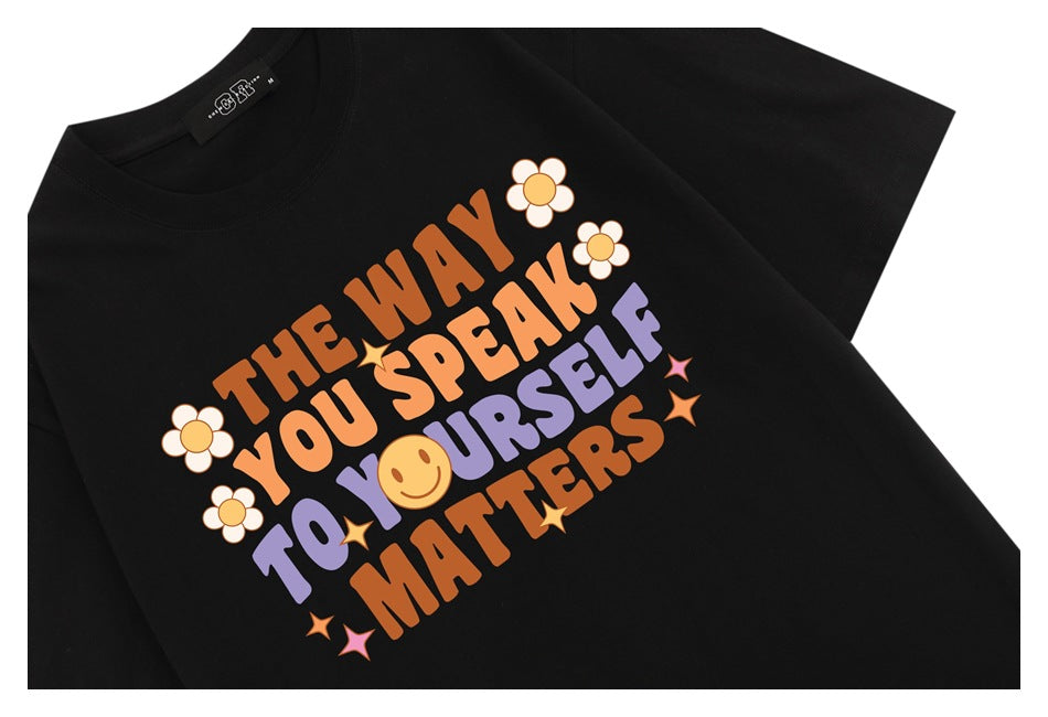 Close view of a black crew-neck T-shirt with orange and lavender lettering, flowers, stars, and a smiley face.
