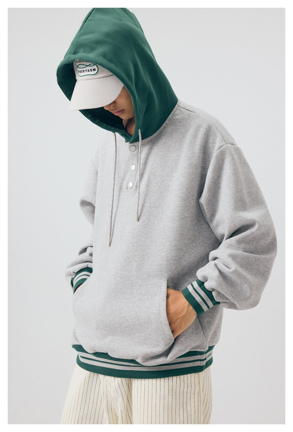Model wearing the gray pullover hoodie with its dark green hood up and hands in the kangaroo pocket.