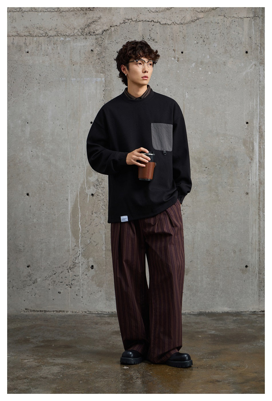 Model wearing a black oversized crewneck sweatshirt with a gray chest pocket and wide striped trousers.
