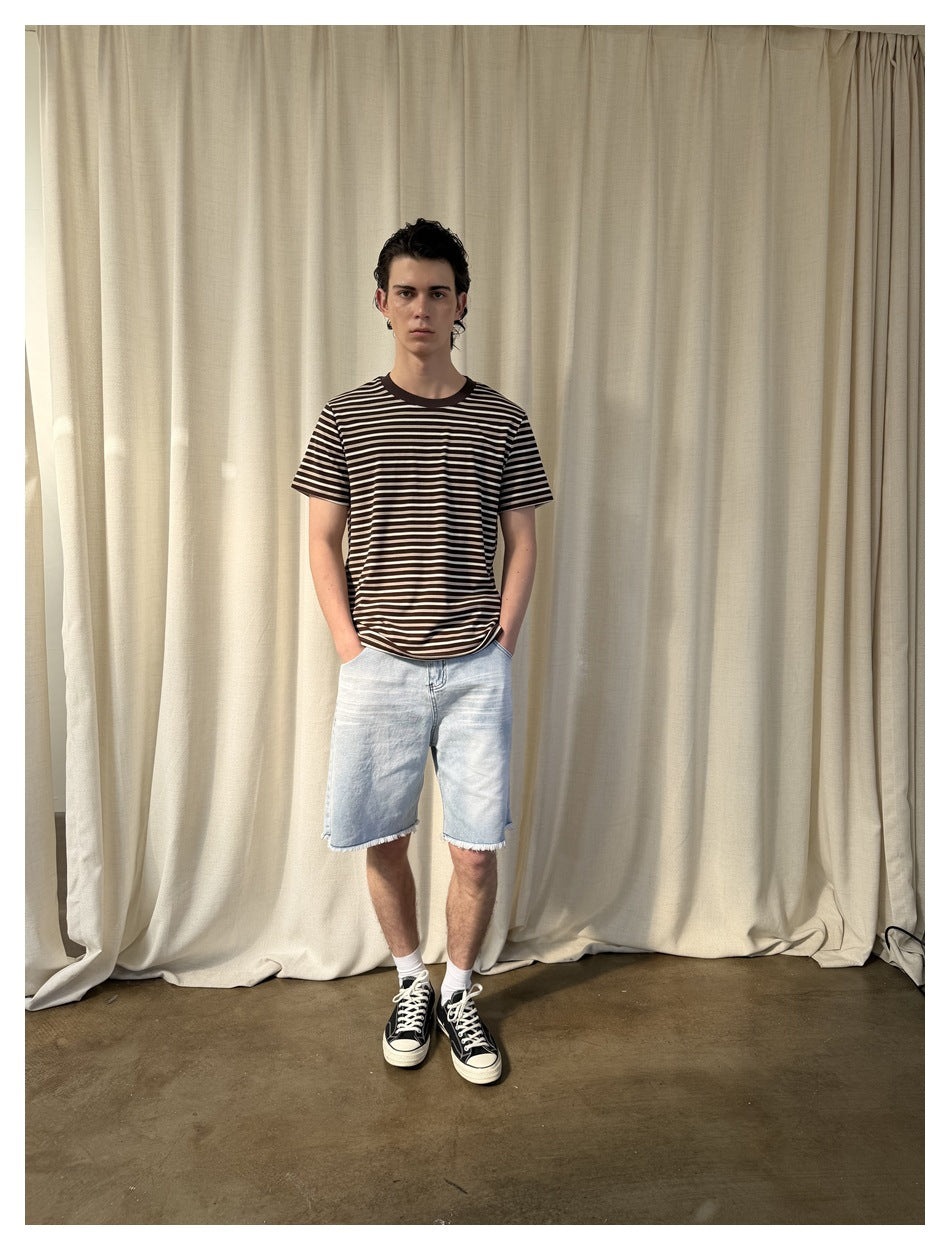 Male model in a brown striped T-shirt, light denim shorts and black sneakers, standing in front of a cream curtain