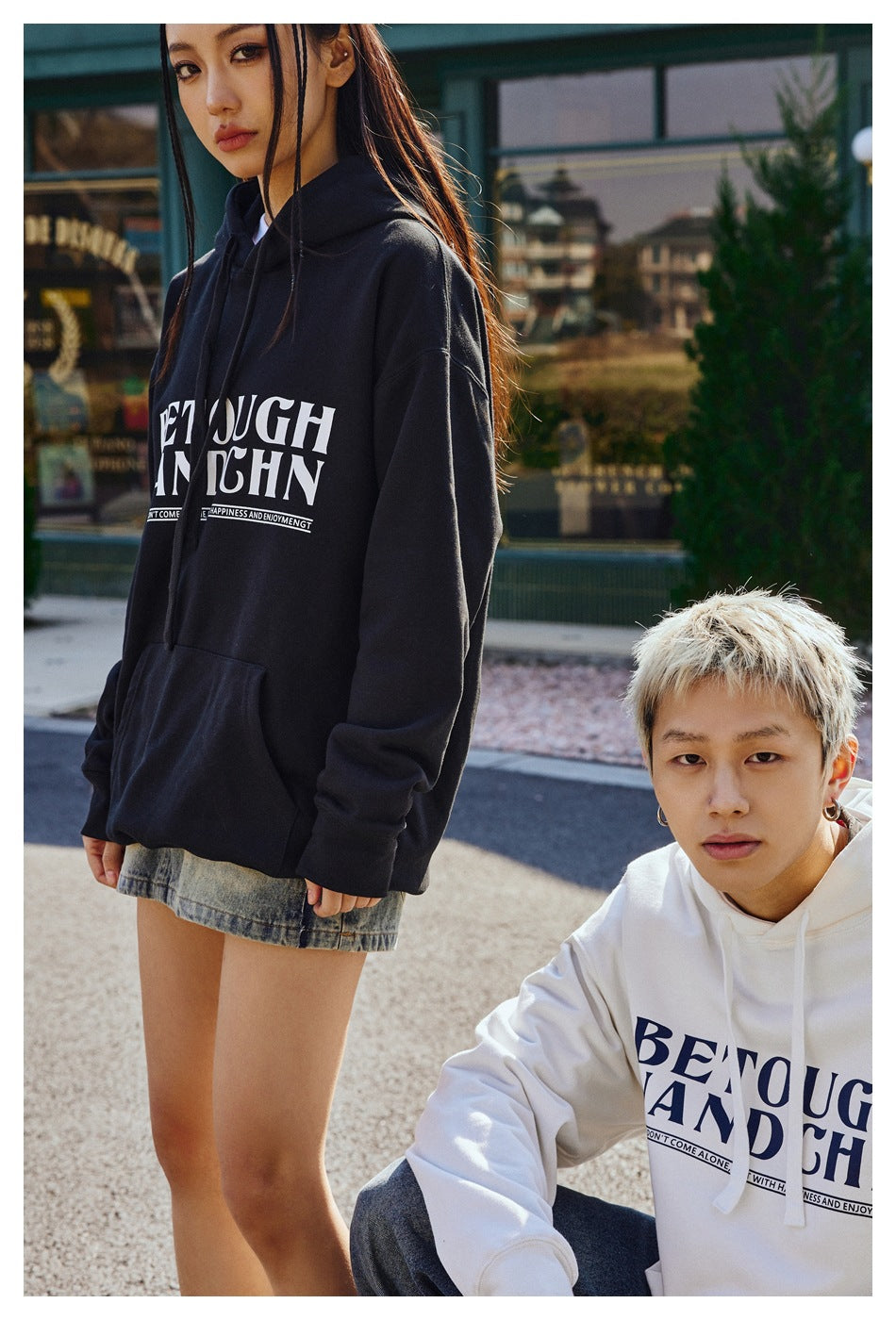 Close view of models wearing black and white graphic hoodies, showing the front lettering, sleeves, and pouch pockets.