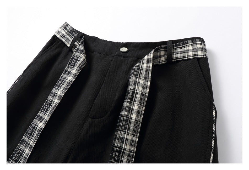 Front close-up of black trousers with plaid waistband accents, hanging plaid ties, and a silver button.