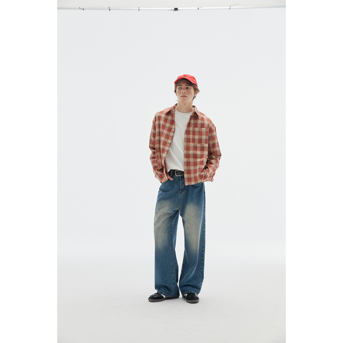 Model wearing a rust-red plaid button-up shirt over a white tee, wide-leg blue jeans, and a red cap.