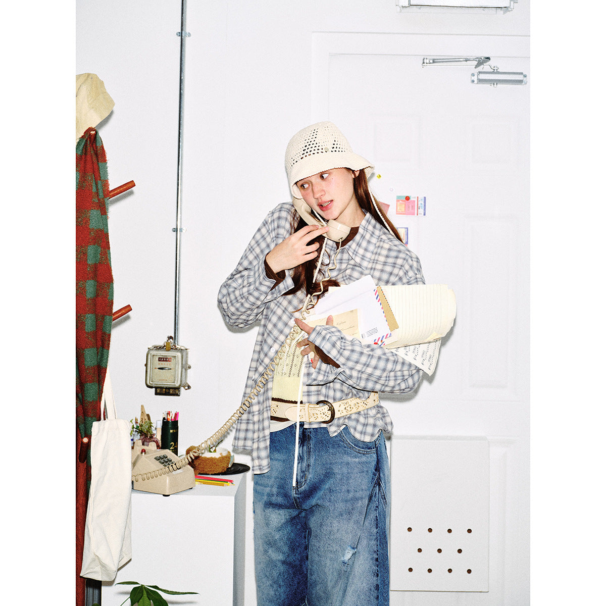 Closer view of a model in a gray-blue plaid shirt, loose blue jeans and cream crochet hat, holding a telephone and papers.