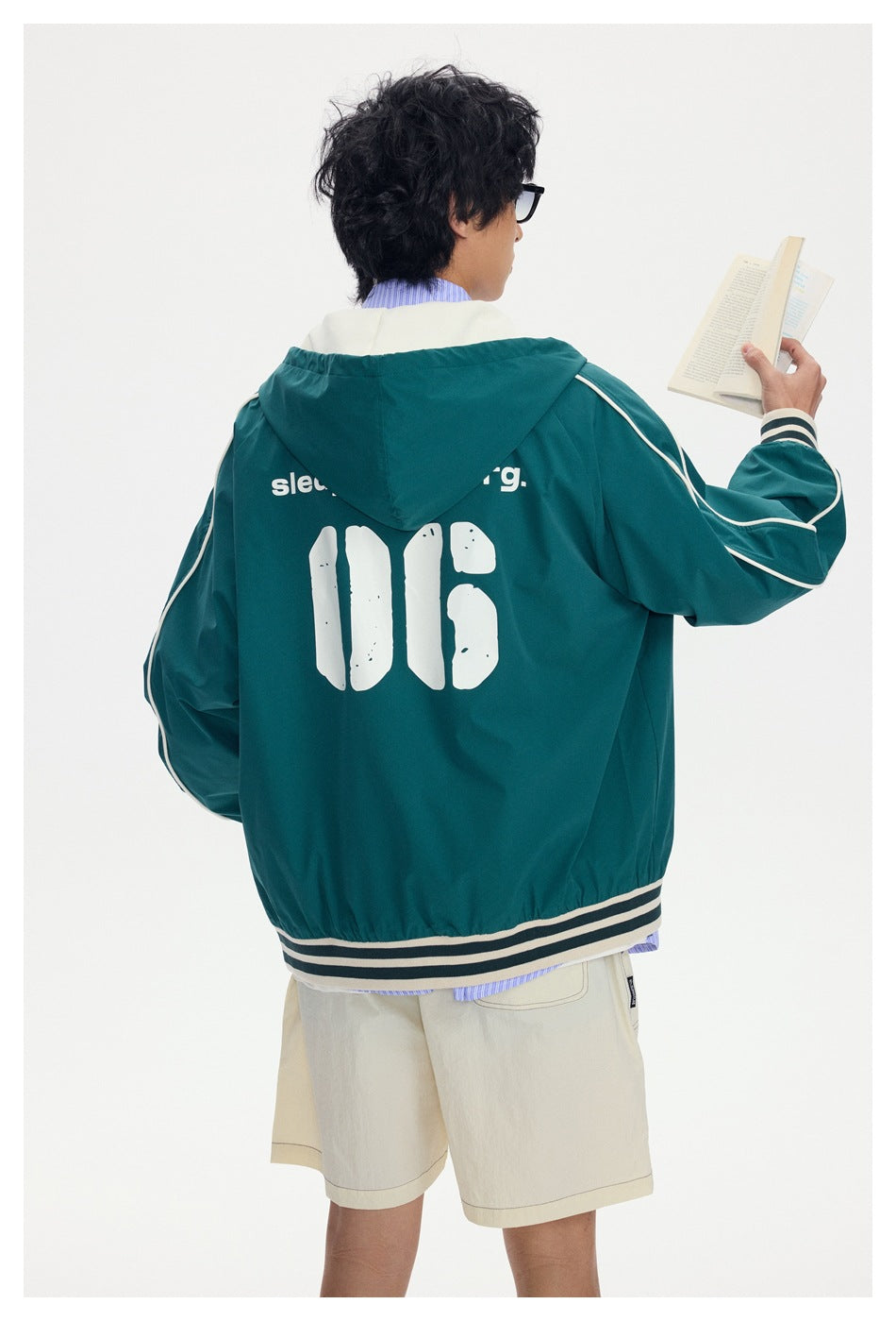 Rear view of a model wearing a teal hooded jacket with cream sleeve piping, striped trim and large white back graphics.