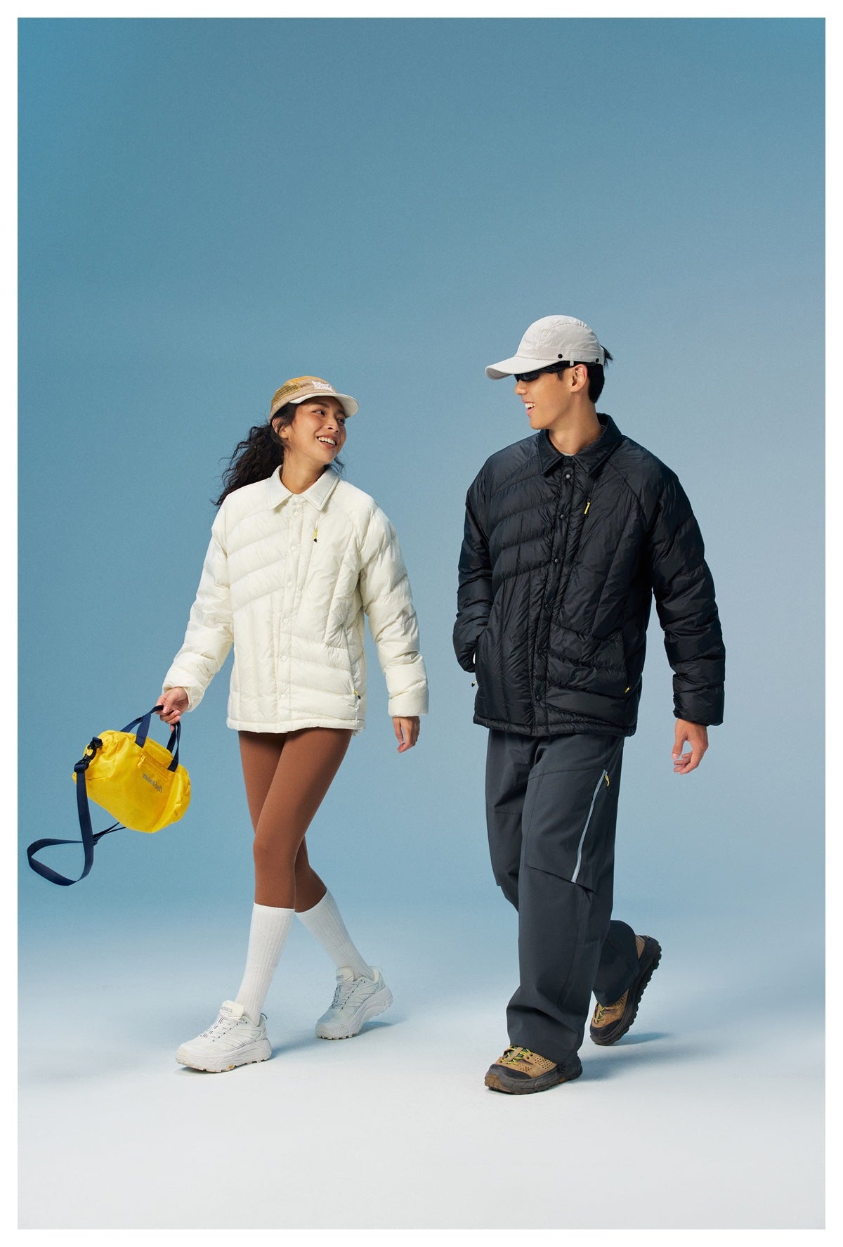 Two models walking in cream and black puffer jackets; the woman carries a yellow bag.