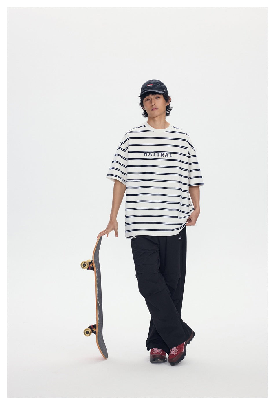 Model wearing an oversized white T-shirt with thin navy stripes and NATURAL text, black pants, and a black cap beside a