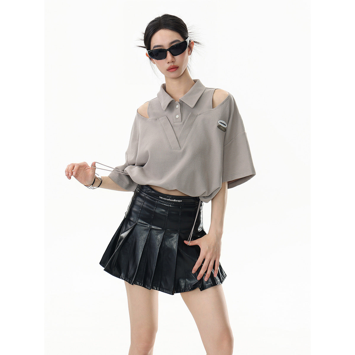 Model wearing a taupe cutout-shoulder polo with a black pleated mini skirt.