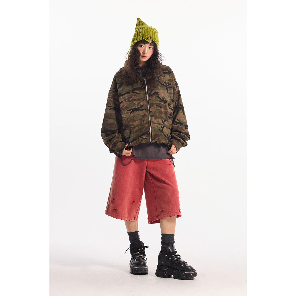 Full-length model wearing a brown and olive camouflage zip jacket with distressed red wide-leg shorts and a lime knit beanie.