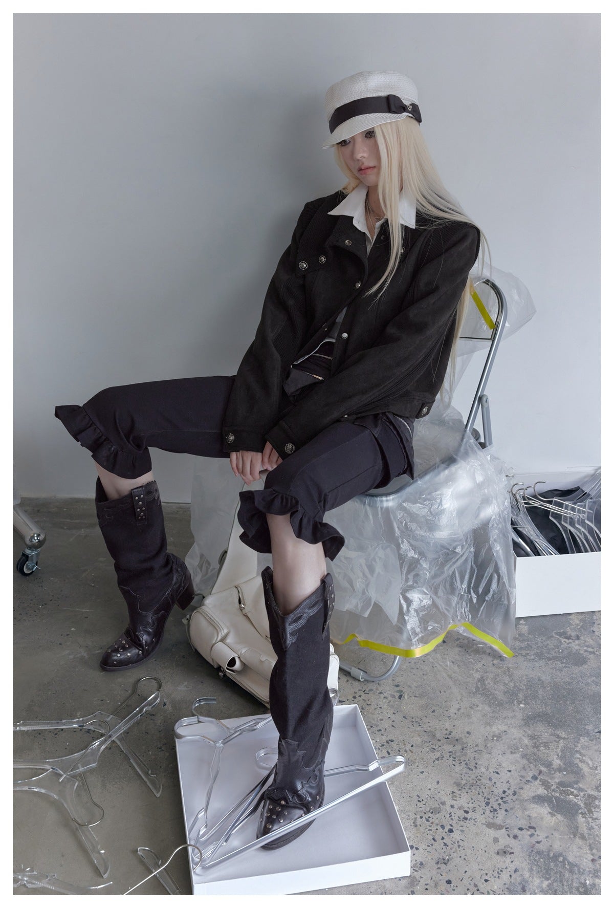 A seated model wears a black buttoned jacket, white shirt, black ruffled pants, tall boots, and a white cap.
