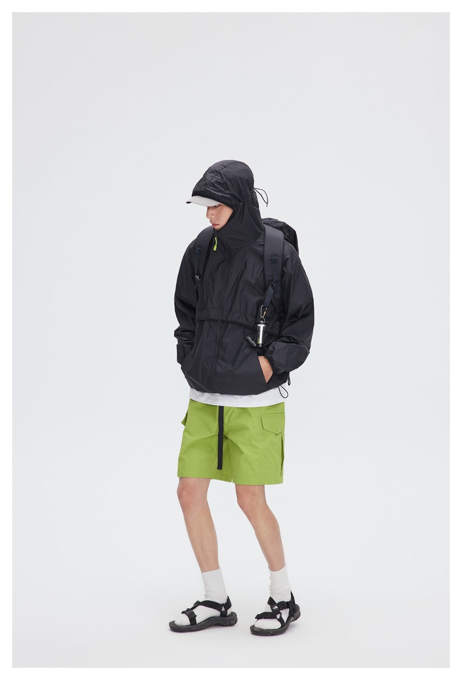 Model wearing a black hooded pullover jacket and lime-green shorts, with the hood up and a backpack.