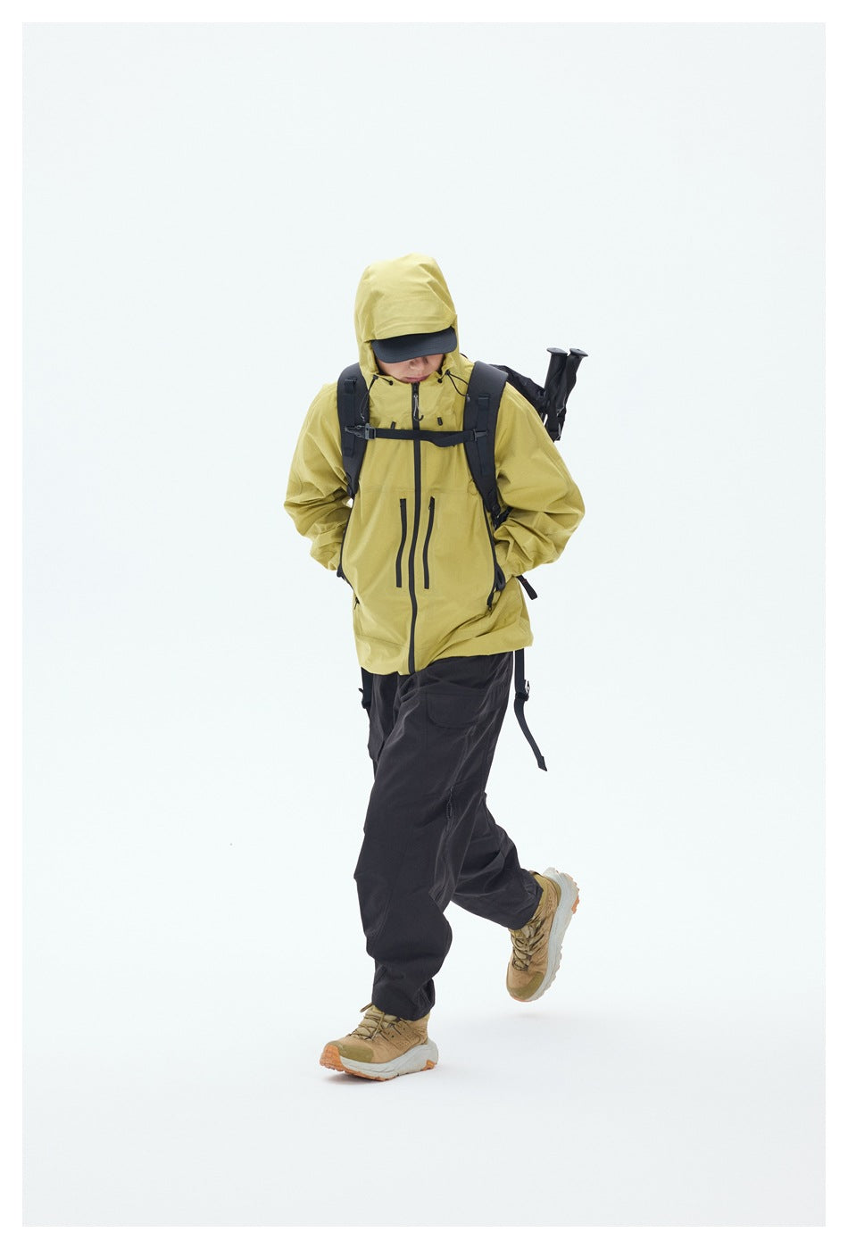 Hiker wears a yellow hooded jacket and black pants with a backpack and trekking poles.