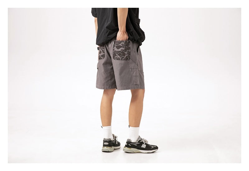 Rear three-quarter view of a model wearing gray shorts with dark camouflage-print pockets.