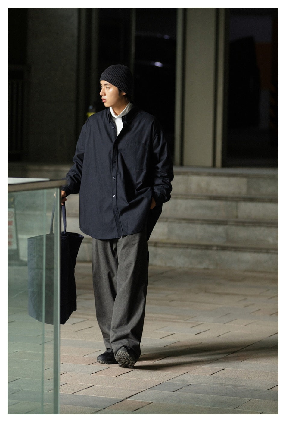 Model wearing an oversized black button-front shirt with a chest pocket, gray wide-leg trousers, and a black beanie.