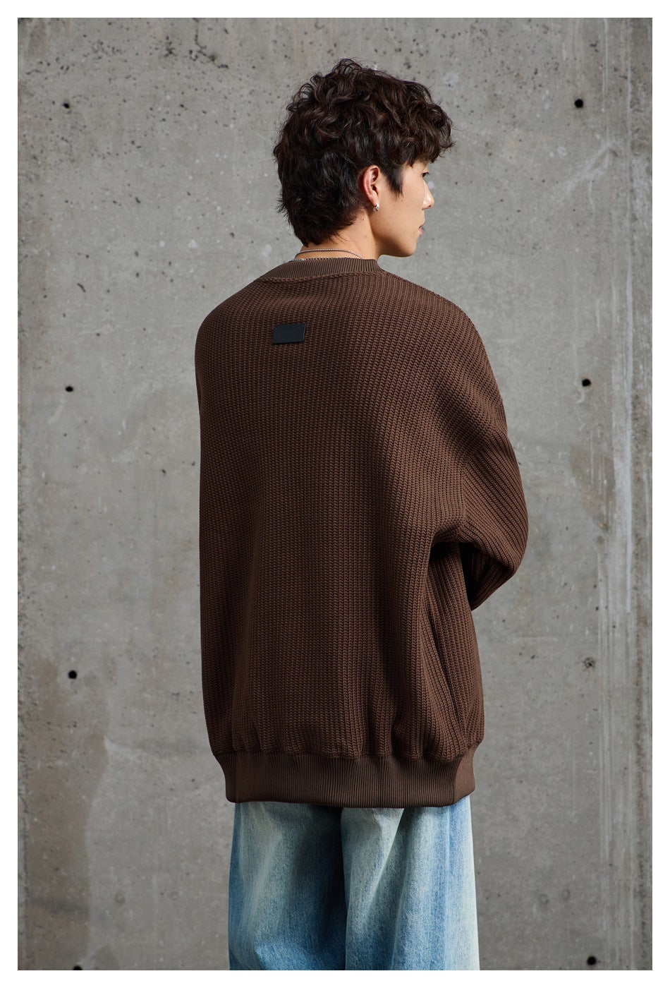 Back view of a model wearing a dark brown textured sweater with ribbed hem and a small black patch near the collar.