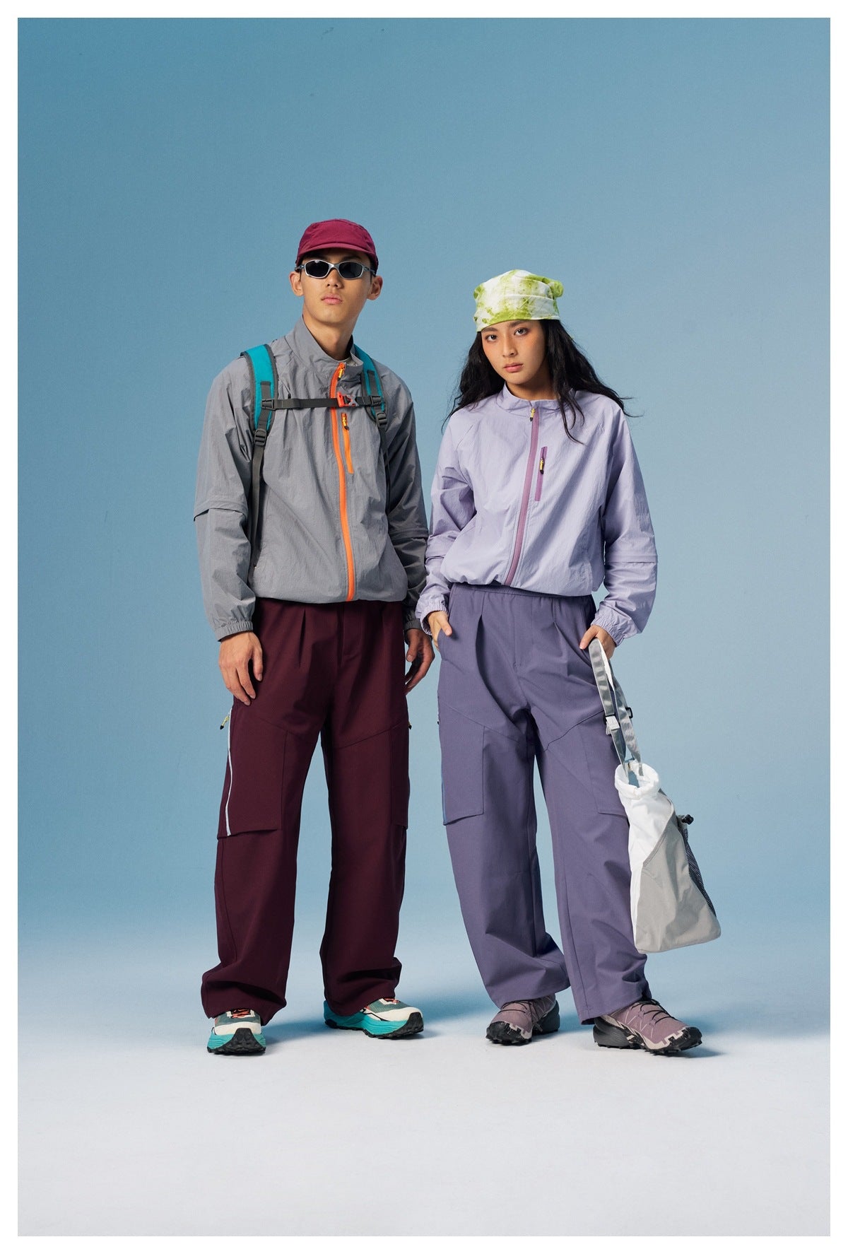 Two models wearing gray and light purple zip jackets with wide-leg pants against a blue studio background