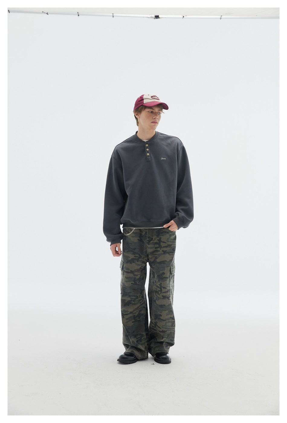 Full-length model wearing a charcoal button-placket sweatshirt with loose camouflage cargo pants and a two-tone cap.