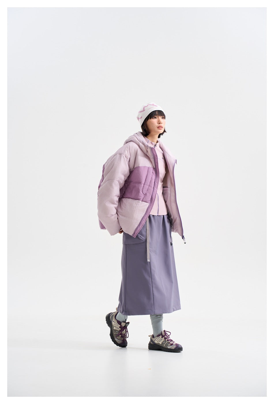 Model in a pale pink and lavender color-block hooded puffer jacket, lavender skirt, and knit hat.
