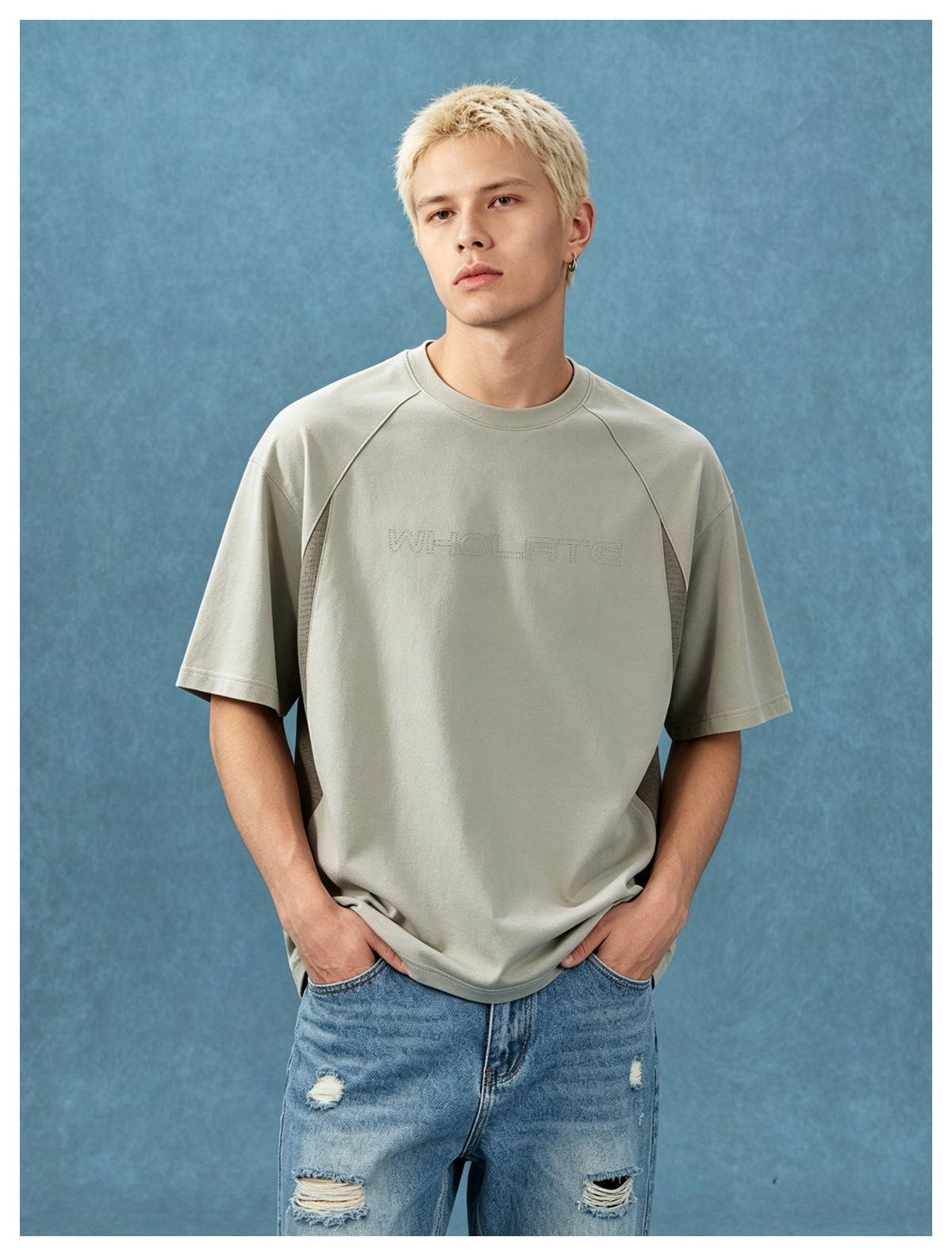 Model wearing a light gray-khaki raglan T-shirt with side panels and ripped light-wash jeans against a blue backdrop
