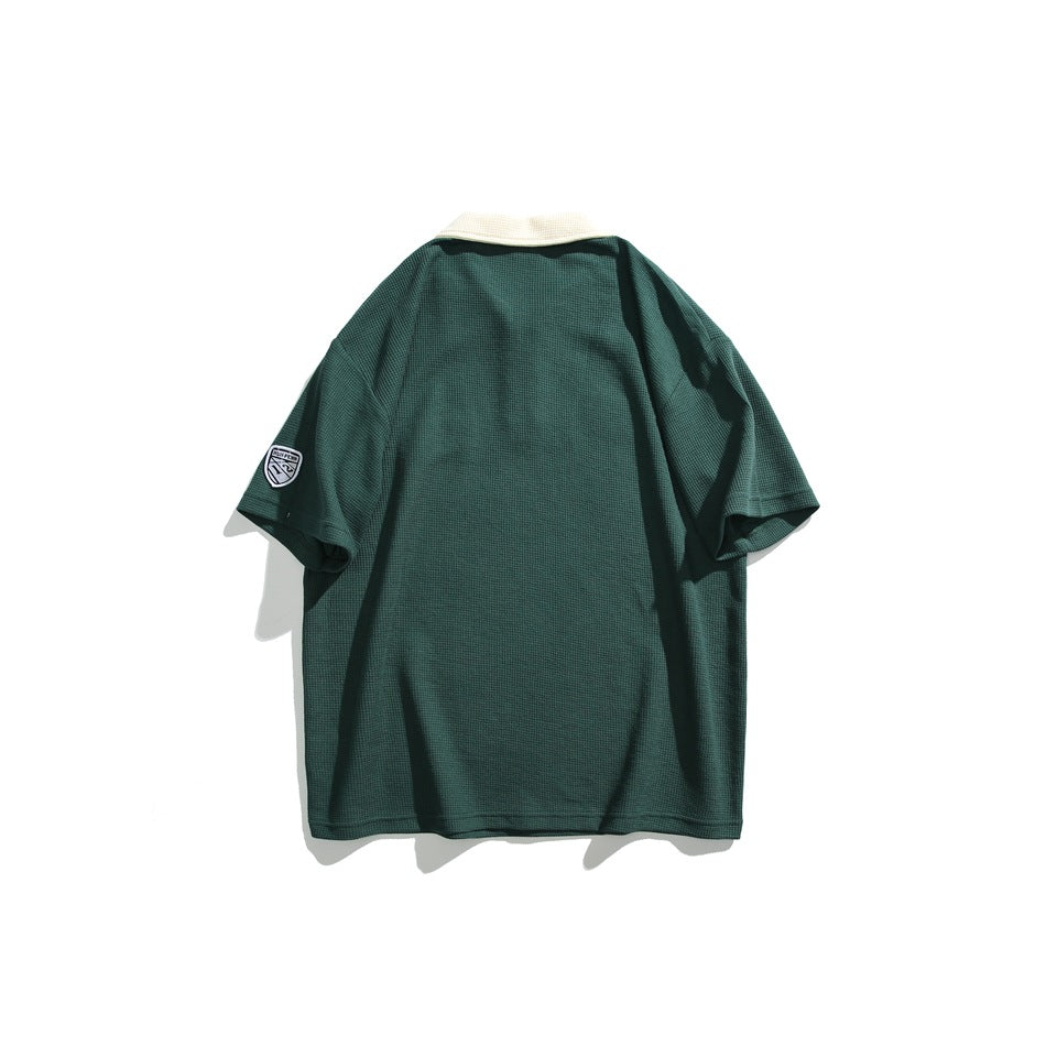 Back view of a dark green short-sleeve polo with an ivory collar and a small shield patch on the sleeve.