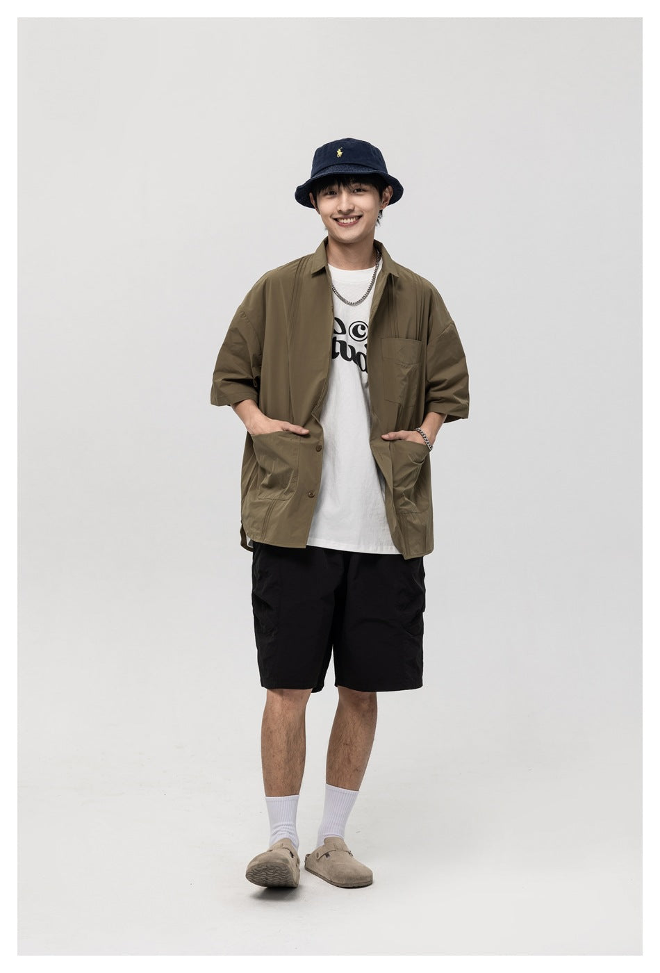 Model wearing green short-sleeve shirt open over a white graphic tee with black shorts and a bucket hat