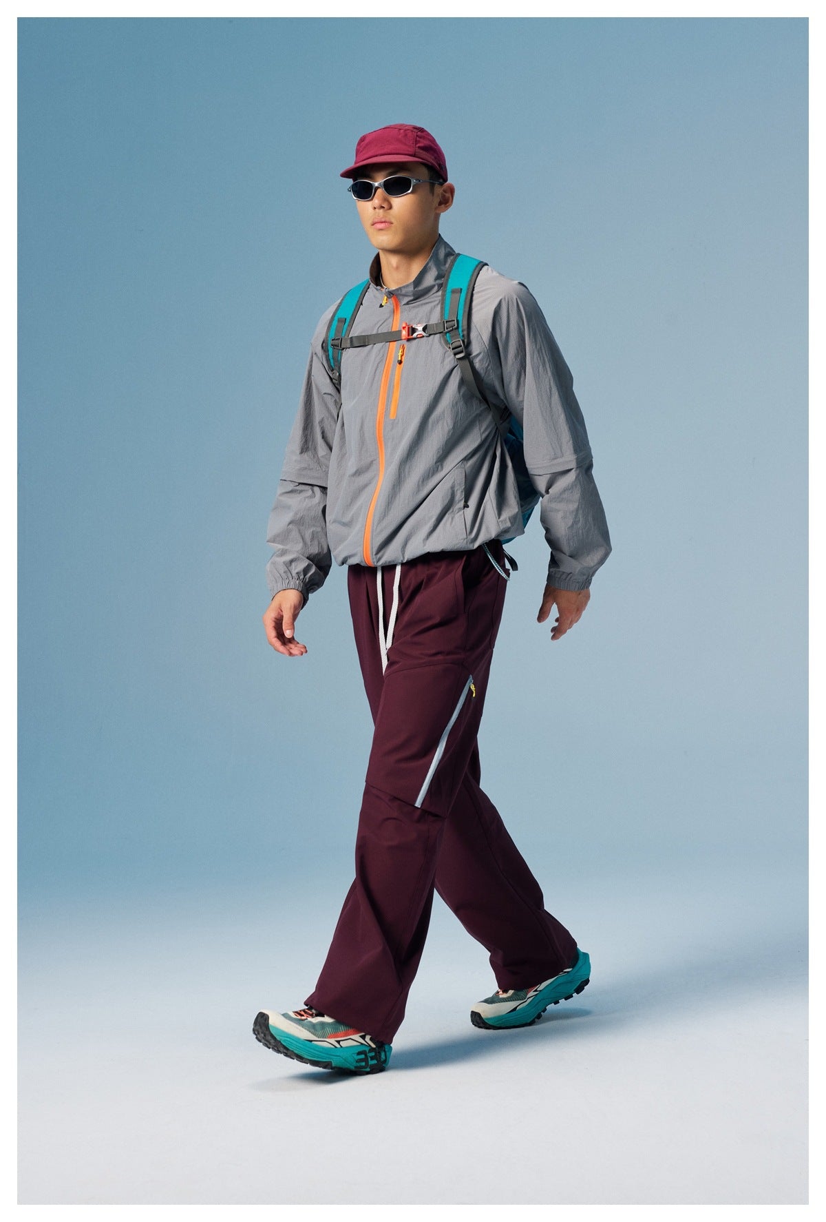 Model walking in a gray orange-zip jacket, burgundy cargo pants, and a teal backpack.