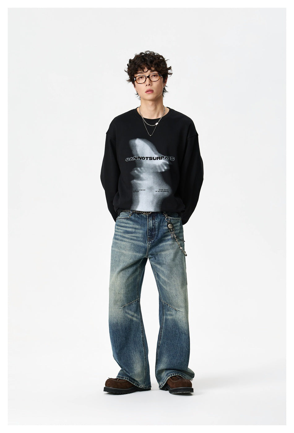 Full-length front view of a model in a black graphic sweatshirt, loose blue jeans, and brown shoes.