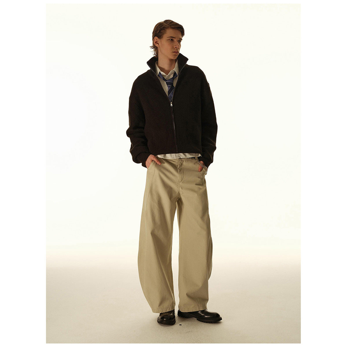 Model wearing a dark zip-up cardigan with a raised collar, striped tie, and loose beige trousers.