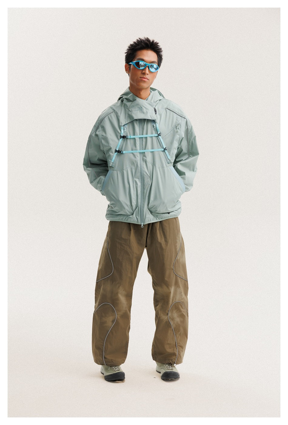 Model wearing a pale sage hooded jacket with turquoise chest straps, loose brown pants and blue sunglasses.