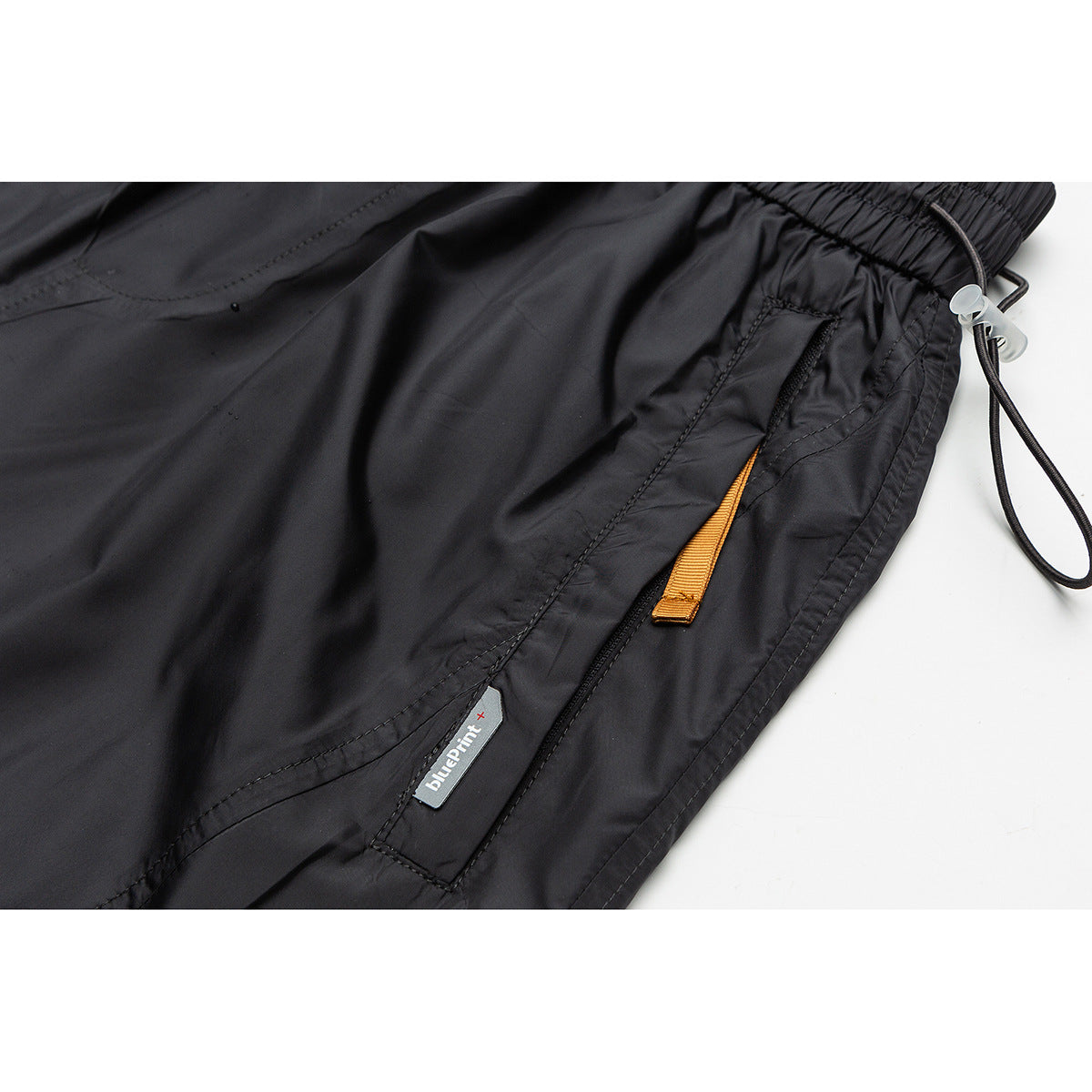 Close-up of black shorts with a zip pocket, orange pull tab, drawcord, and a small attached brand label.