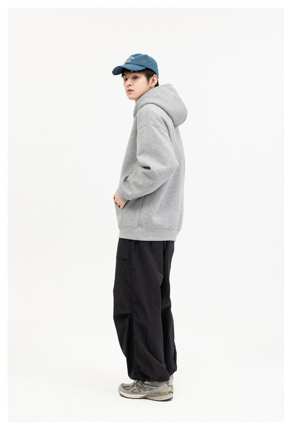 Model in a gray hoodie shown from the side, with black pants and a blue embroidered cap.