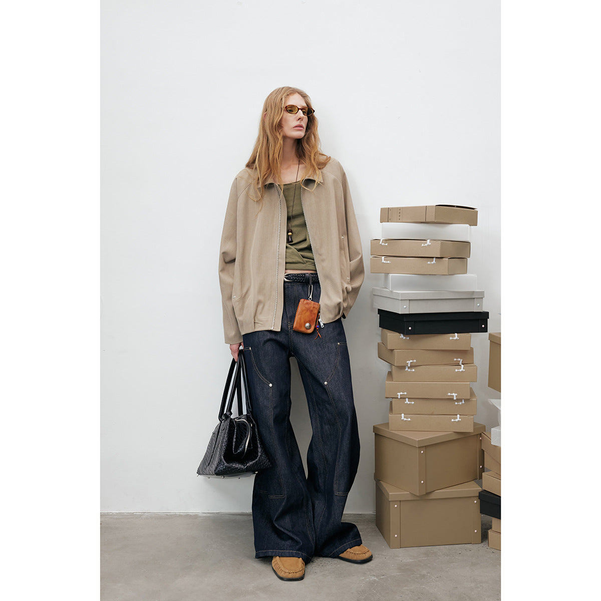 Model wears a taupe zip-front jacket over an olive top and wide dark jeans, standing beside stacked boxes.