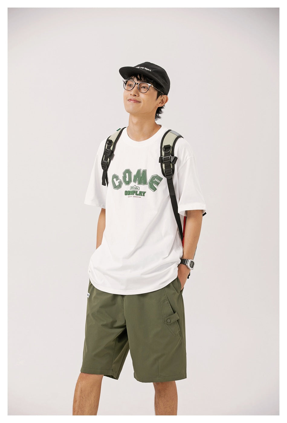 Model wears a white graphic T-shirt with olive-green cargo shorts and a backpack.