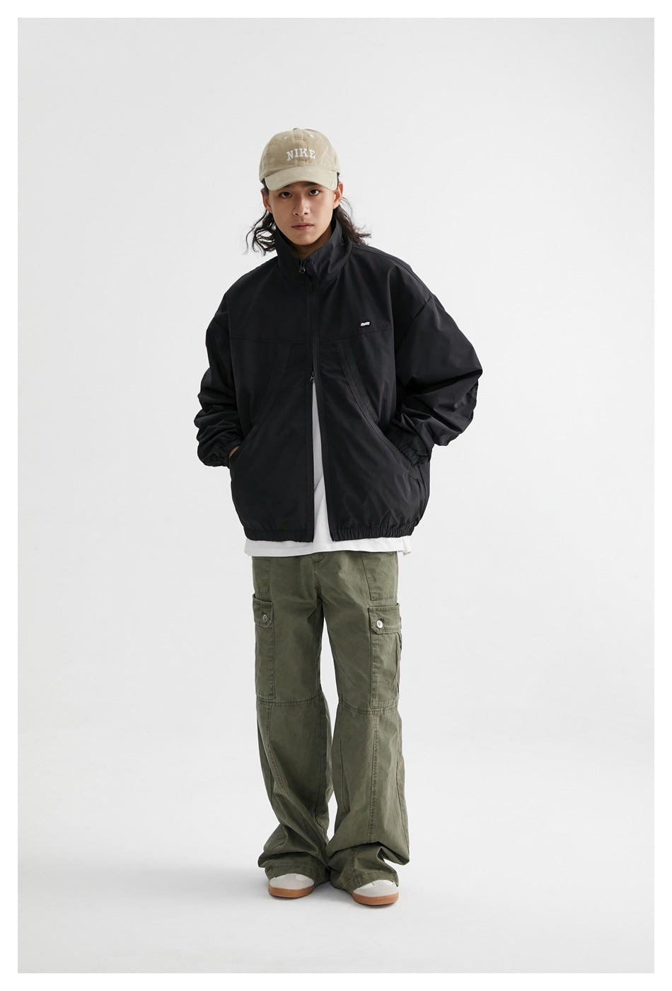 Model wearing the black zip jacket over a white tee with olive cargo pants and a beige cap, hands in pockets