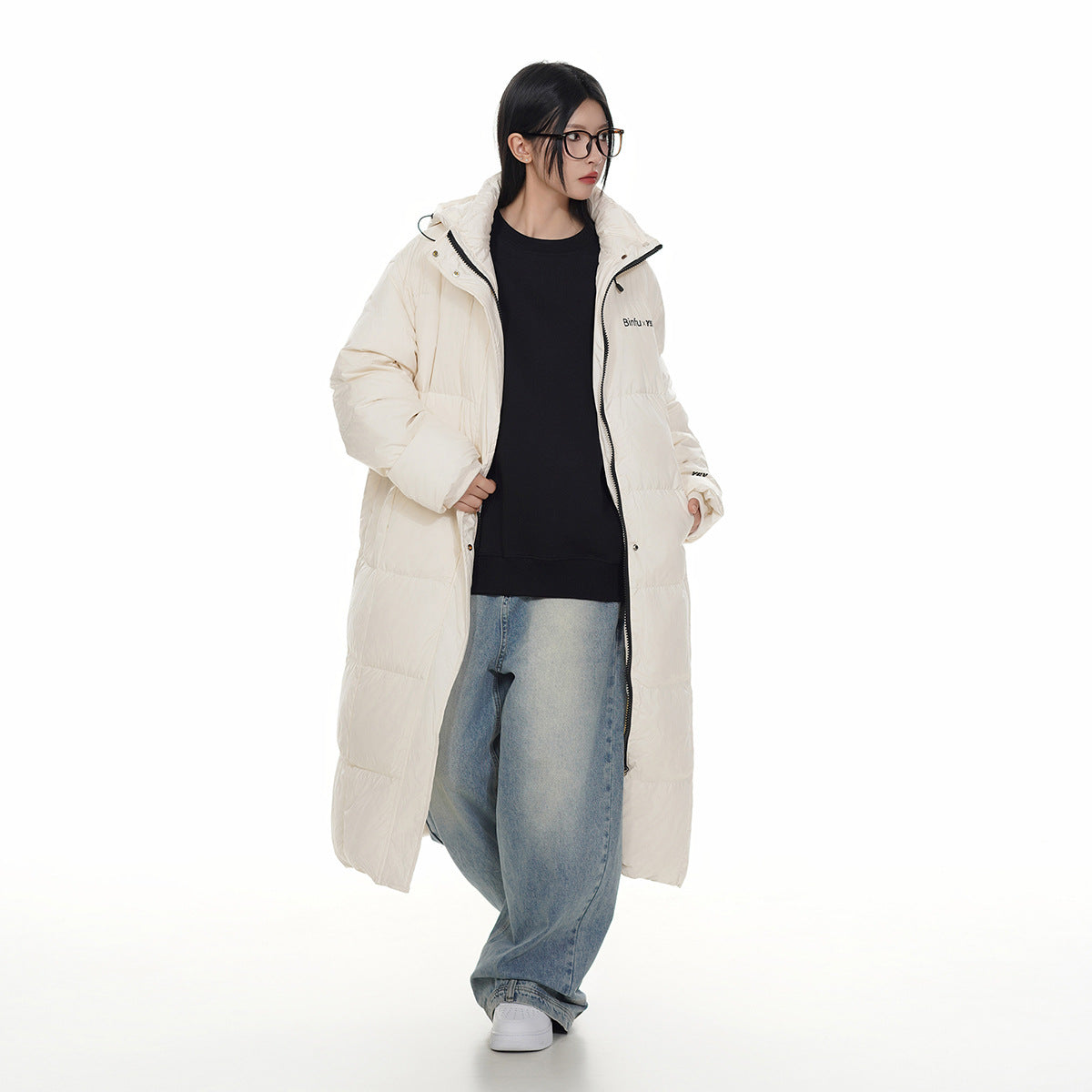 Model wearing a long ivory hooded puffer coat open over a black top and loose blue jeans.