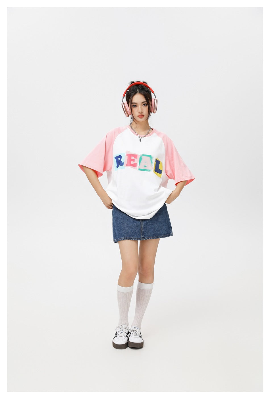 Model wearing a white raglan T-shirt with pink sleeves and colorful REAL lettering, paired with a denim skirt.