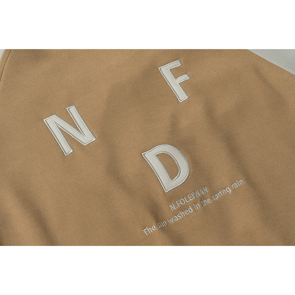 Close-up of tan jacket fabric with raised white N, F, and D lettering and a small embroidered English phrase.