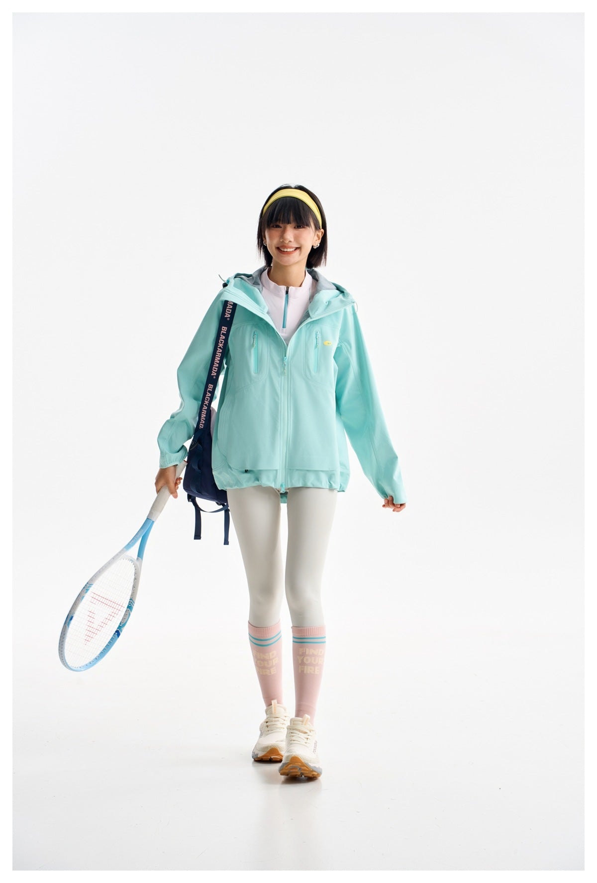Model wearing a mint turquoise hooded jacket with zip chest pockets, holding a tennis racket and wearing a backpack.