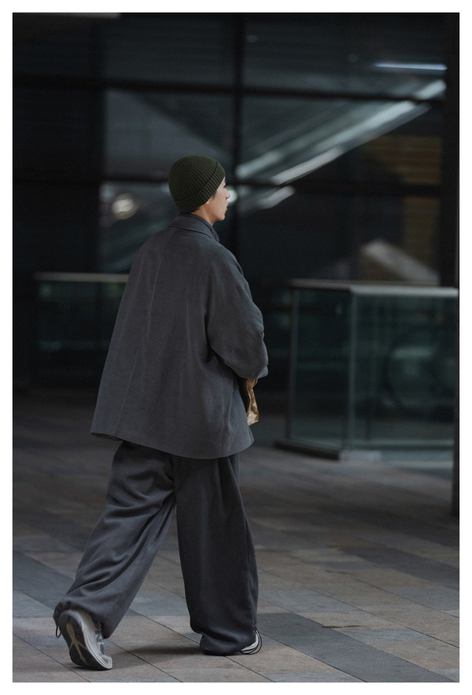 Rear view of a model wearing a dark gray oversized coat and wide-leg pants with an olive knit beanie.